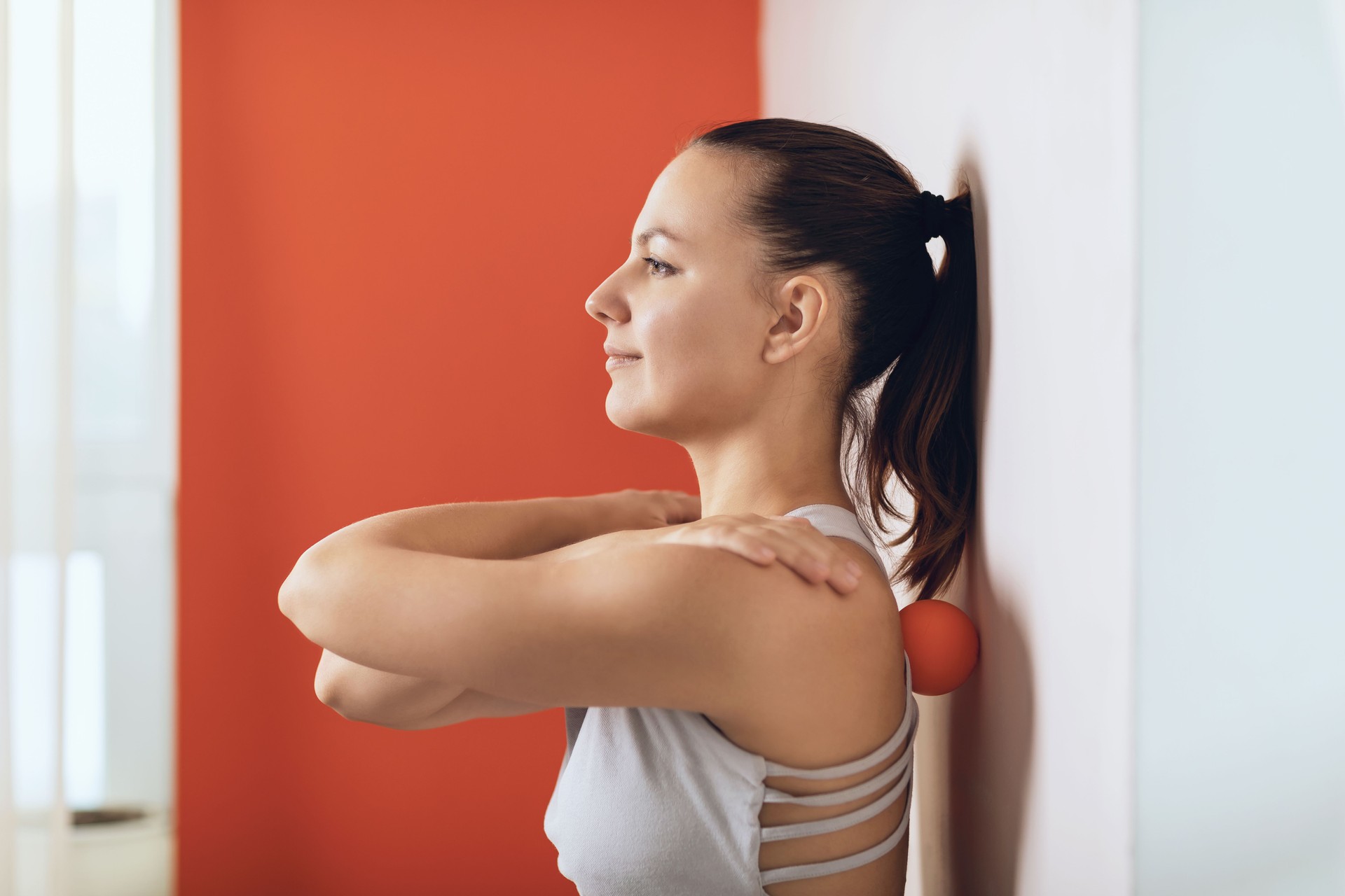 Beautiful woman performs myofascial release of hyper-flexible muscles of the back with a massage ball near the wall. The concept of back fatigue prevention and self-massage